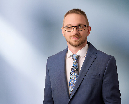 A man, Keith Brown, in a blue suit, white shirt, and patterned blue tie is looking at the camera, standing against a blurred blue and white background.